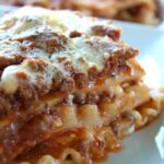 A close-up shot of a slice of classic homemade meat lasagna on a white plate, showing layers of melted mozzarella, rich meat sauce, and wavy pasta.