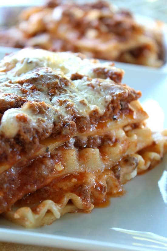A close-up shot of a slice of classic homemade meat lasagna on a white plate, showing layers of melted mozzarella, rich meat sauce, and wavy pasta.