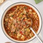 Homemade cabbage roll soup simmering in a white Dutch oven with a ladle, topped with grated Parmesan cheese and chopped parsley, served with crusty bread.