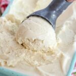 A hand scooping creamy old-fashioned homemade vanilla ice cream with a metal scoop from a light blue container, showing rich texture and vanilla bean flecks.