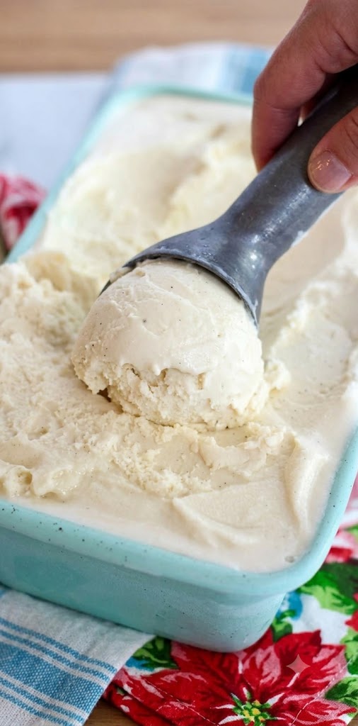 A hand scooping creamy old-fashioned homemade vanilla ice cream with a metal scoop from a light blue container, showing rich texture and vanilla bean flecks.