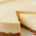 A full round no-bake vanilla bean cheesecake with a slice removed, showing a creamy rich filling and buttery graham cracker crust on a white plate.